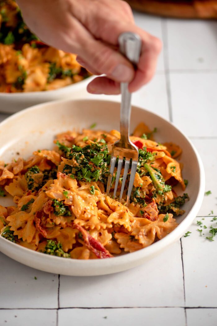 Hand using a fork in a bowl of creamy vegan sun dried tomato and kale bowtie pasta.