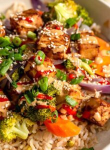 Overhead view of a colourful bowl with tofu, broccoli, carrot, onion and rice, topped with creamy peanut sauce, green onion, sesame seeds and hot sauce.
