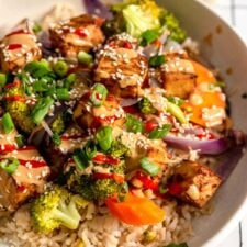 Overhead view of a colourful bowl with tofu, broccoli, carrot, onion and rice, topped with creamy peanut sauce, green onion, sesame seeds and hot sauce.