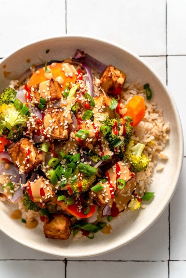 Overhead view of a colourful bowl with tofu, broccoli, carrot, onion and rice, topped with creamy peanut sauce, green onion, sesame seeds and hot sauce.