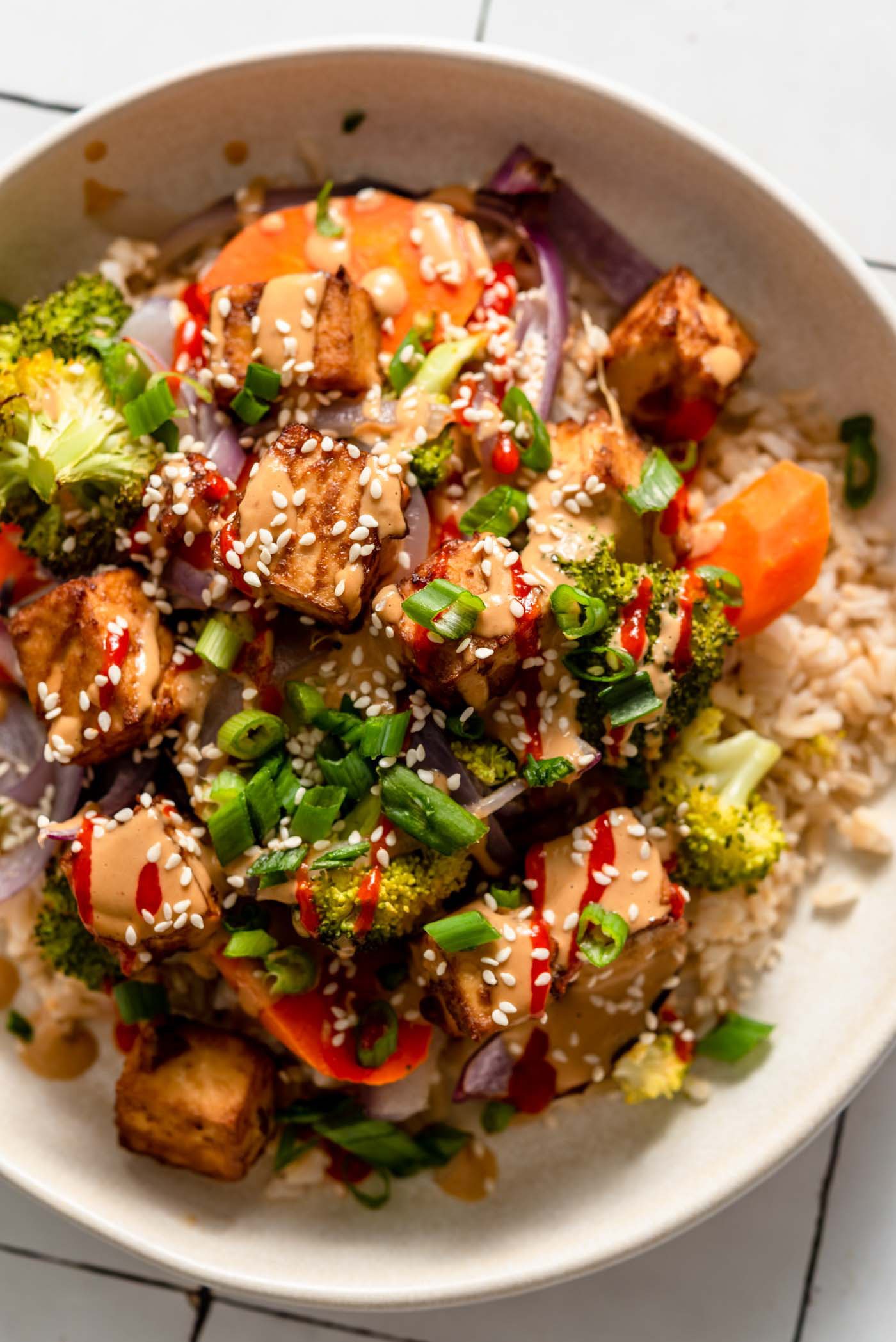 Overhead view of a colourful bowl with tofu, broccoli, carrot, onion and rice, topped with creamy peanut sauce, green onion, sesame seeds and hot sauce.