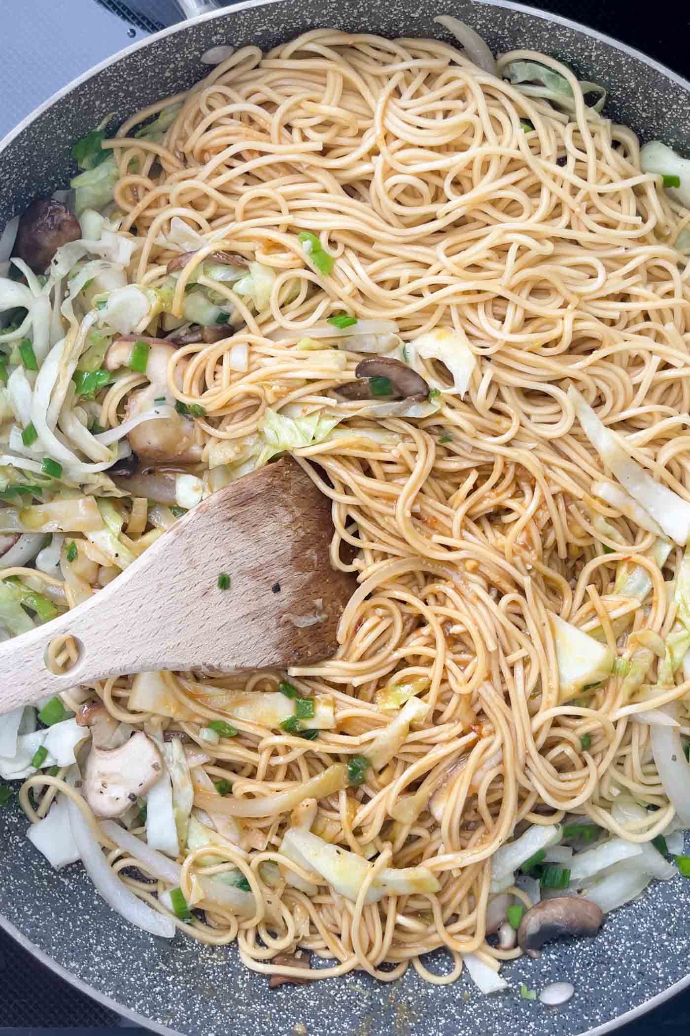 Noodles and sauce with cabbage and mushroom cooking in a large skillet with a wooden spoon.