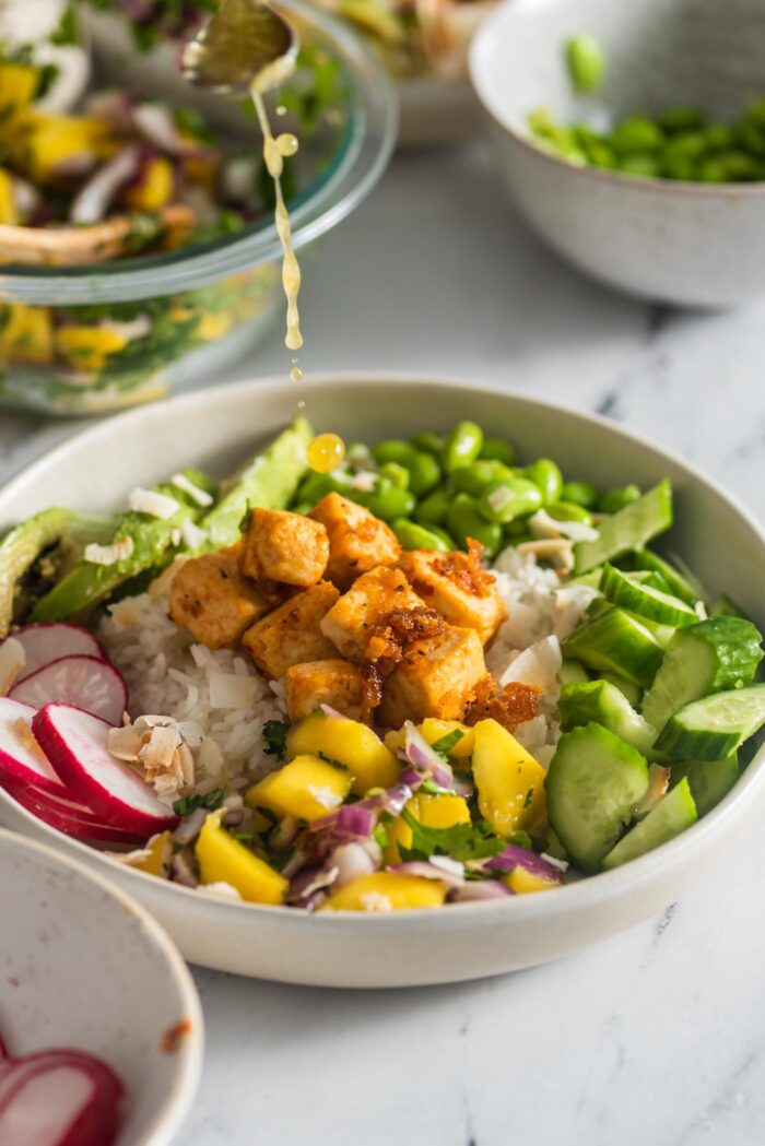 Drizzling ginger lime dressing from a small spoon over a colourful bowl with rice, mango, edamame, avocado and radish.