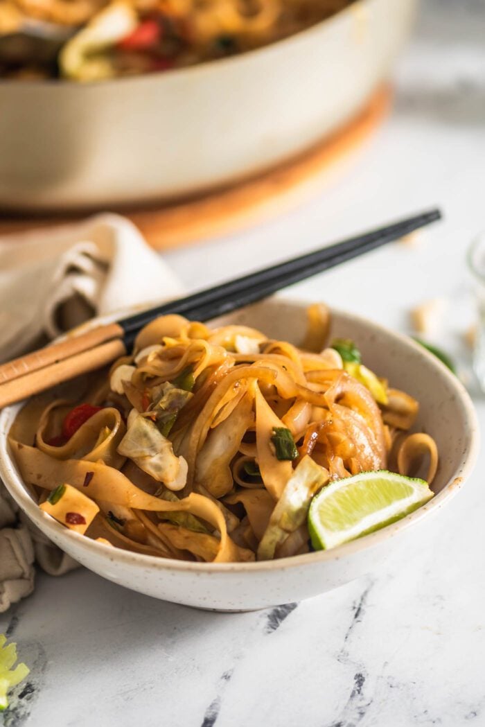 Pair of chopsticks resting on a bowl of vegan drunken noodles with a wedge of lime resting in the bowl.