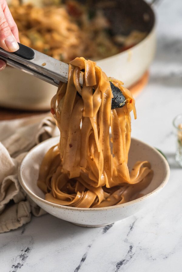 Using tongs to scoop a serving of Thai drunken noodles into a bowl.