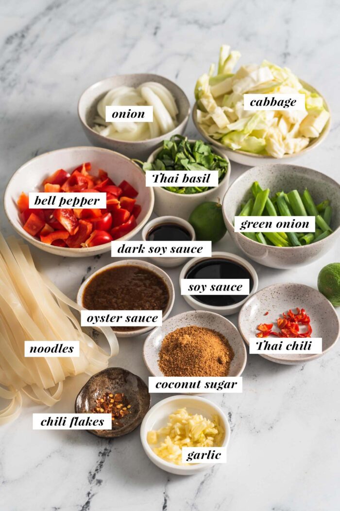 All the ingredients needed for a pad kee mao recipe gathered in small bowls. Each ingredient is labelled with text.