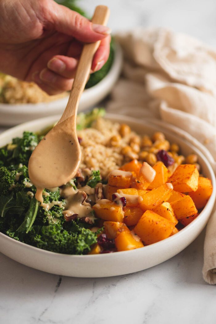 Hand using a spoon to drizzle dressing over a healthy bowl of kale, quinoa and squash.