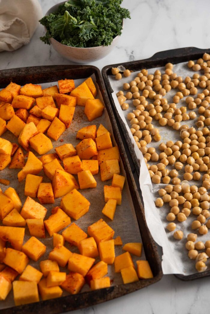 One baking pan with cubed butternut squash on it beside another with chickpeas on it. Each pan is lined with parchment paper.