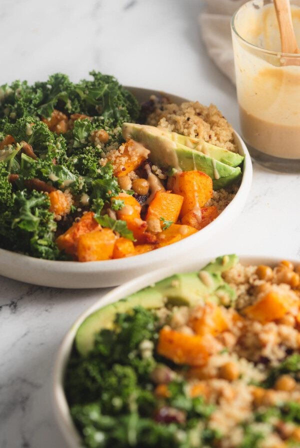 Two quinoa power bowls with butternut squash, chickpeas, kale and avocado. A jar of maple dijon dressing is in the background.