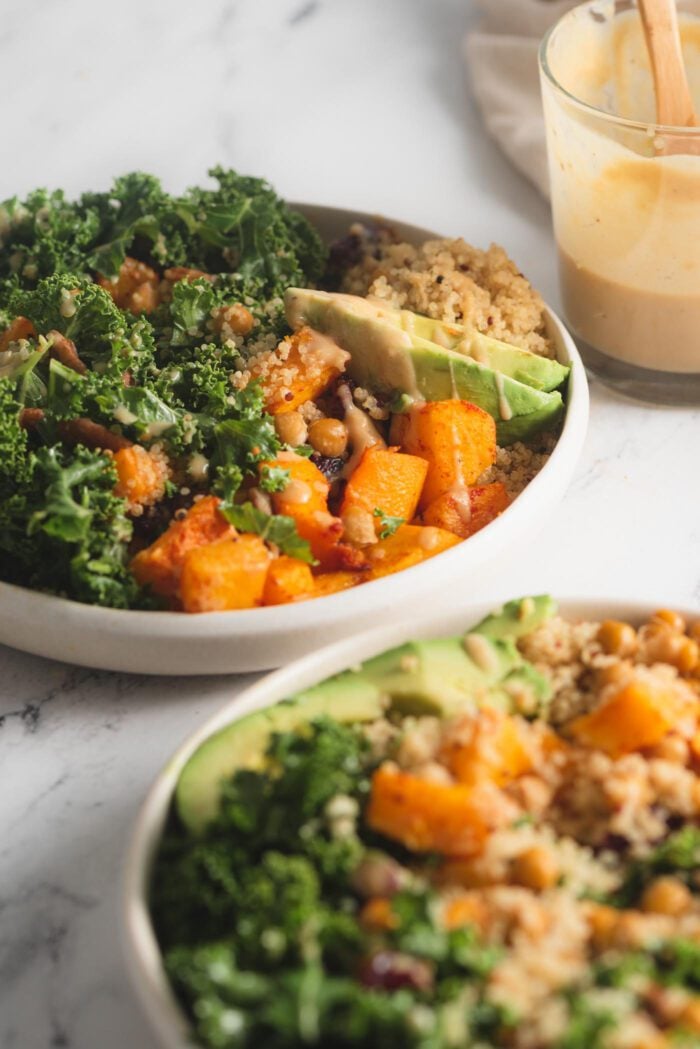 Two quinoa power bowls with butternut squash, chickpeas, kale and avocado. A jar of maple dijon dressing is in the background.