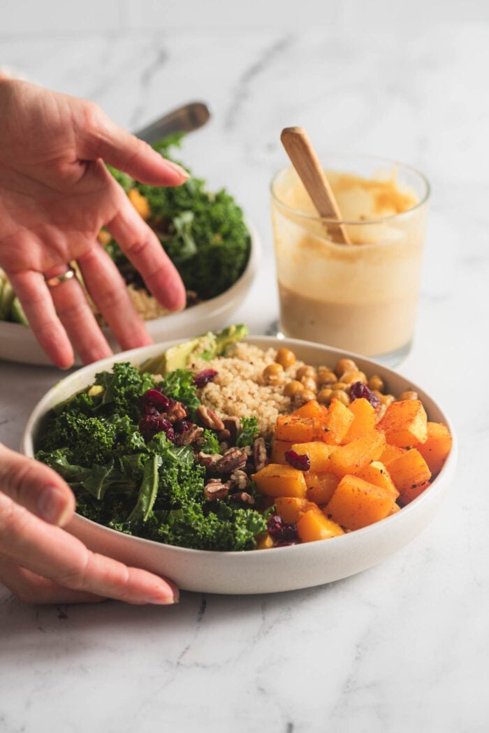 Hands placing a quinoa, squash, kale and cranberry bowl on a counter. A small jar of dressing is in the background.