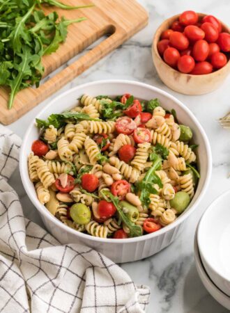 A bowl of zesty Italian pasta salad with tomato, artichokes, arugula and olives. A bowl of tomatoes is in the background.