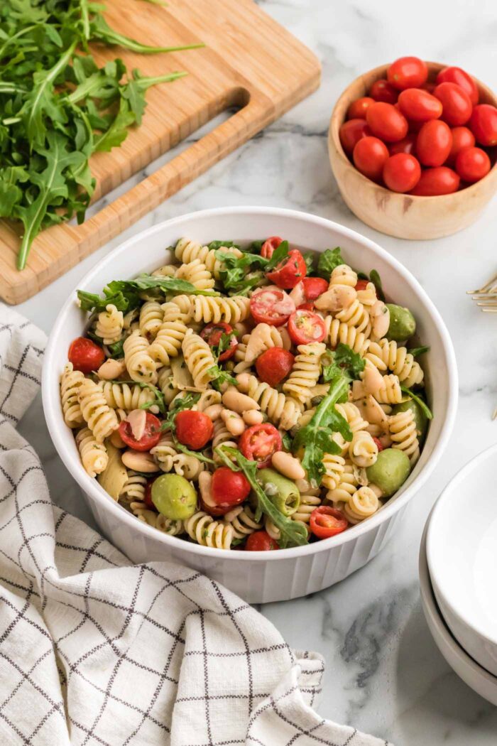 A bowl of zesty Italian pasta salad with tomato, artichokes, arugula and olives. A bowl of tomatoes is in the background.