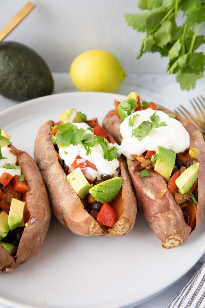Three black bean loaded sweet potatoes topped with sour cream and avocado on a small plate.