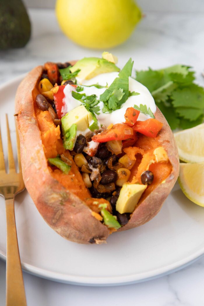 A baked sweet potato sliced open and stuffed with a black bean, red pepper and onion filling and topped with yogurt, avocado and cilantro.