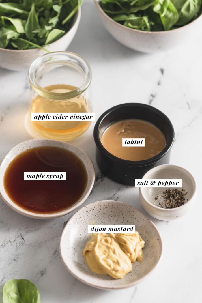Dijon mustard, maple syrup, tahini, apple cider vinegar and salt and pepper in small bowls. Each ingredient is labelled with text.