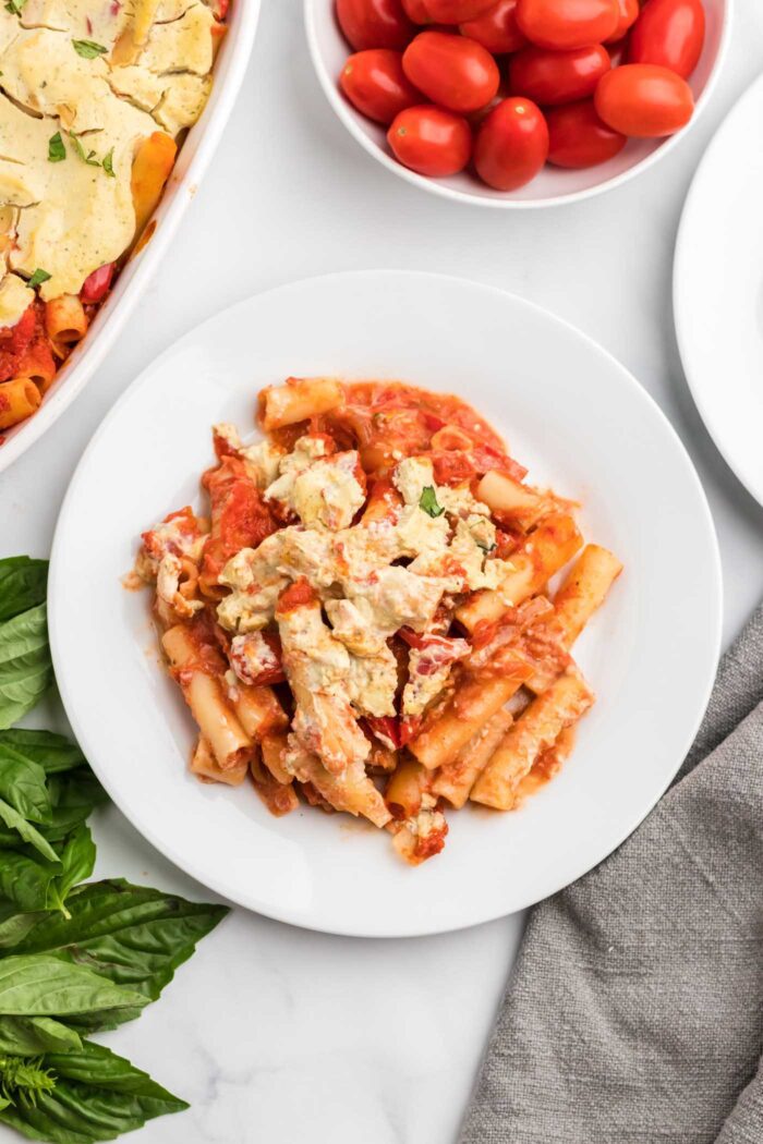 Overhead view of a serving of baked ziti made with tofu ricotta cheese on a small plate. There's a small dish or cherry tomatoes beside the plate and a baking dish of baked ziti.