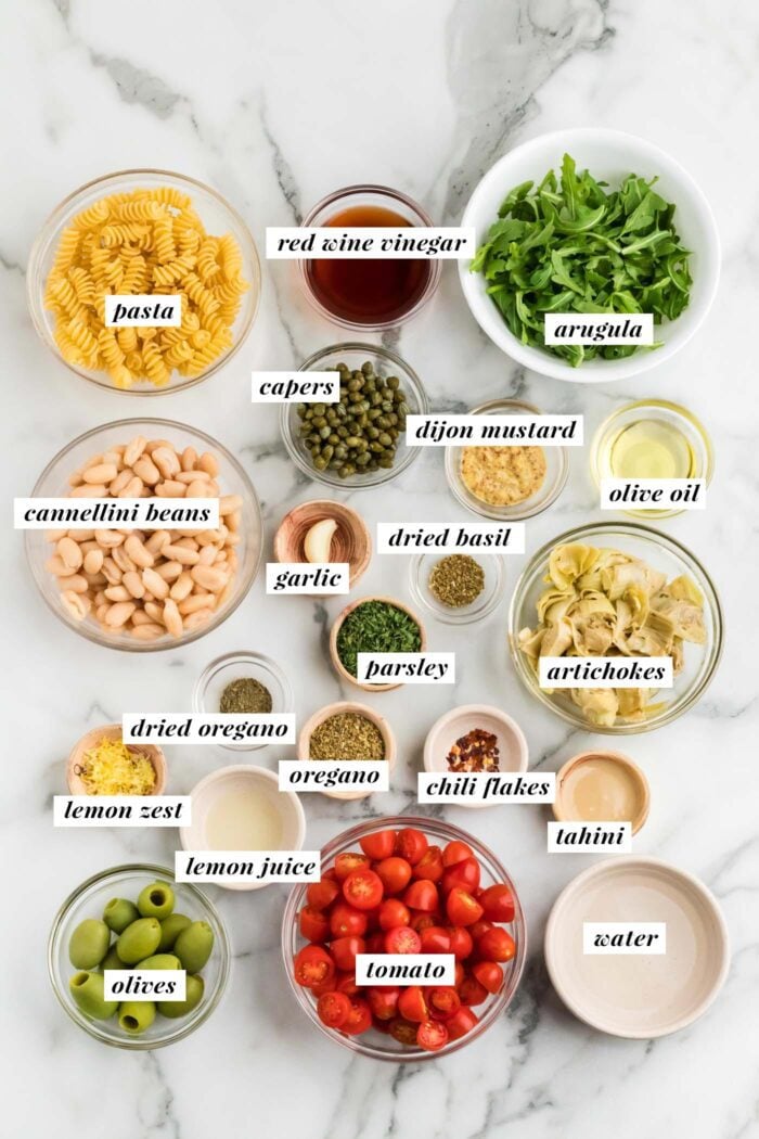 Overhead view of all the ingredients needed for making a vegan zesty Italian pasta salad recipe. Each ingredient is labelled with text.