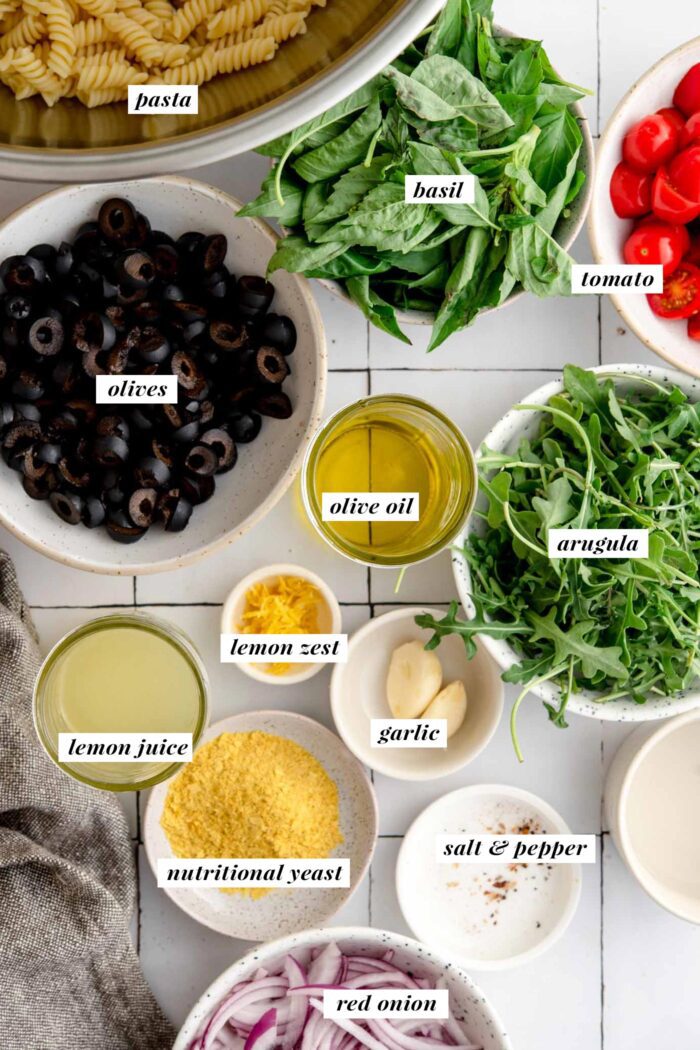 Overhead view of the ingredients needed for making a cold pesto pasta salad recipe with olives, red onion, basil, arugula and tomato.