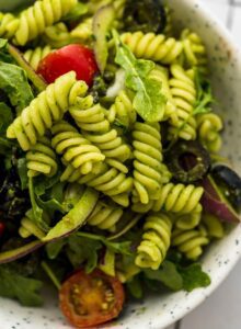 Bowl of cold pesto pasta salad with tomato, arugula, olives and red onion.