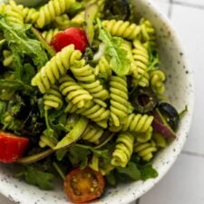 Bowl of cold pesto pasta salad with tomato, arugula, olives and red onion.