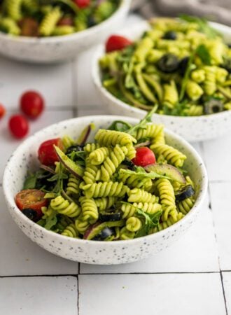 Bowl of cold pesto pasta salad with tomato, arugula, olives and red onion.