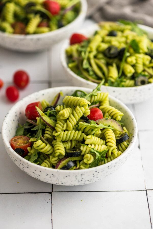 Bowl of cold pesto pasta salad with tomato, arugula, olives and red onion.