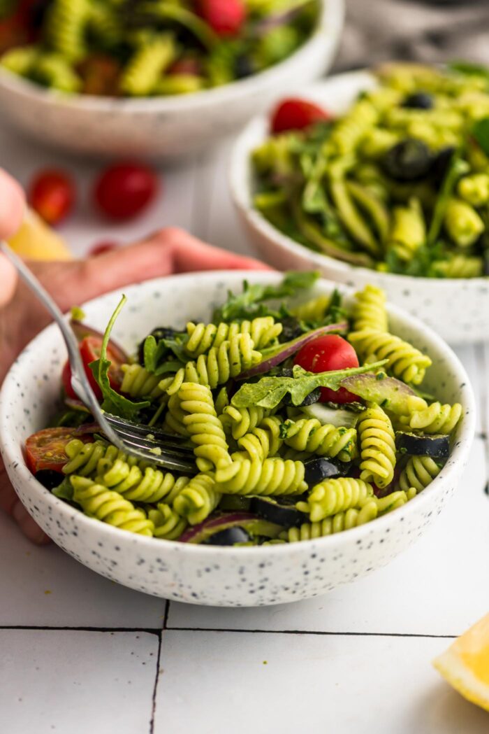 Using a fork in a small bowl of pesto pasta salad with tomato, arugula and olives.