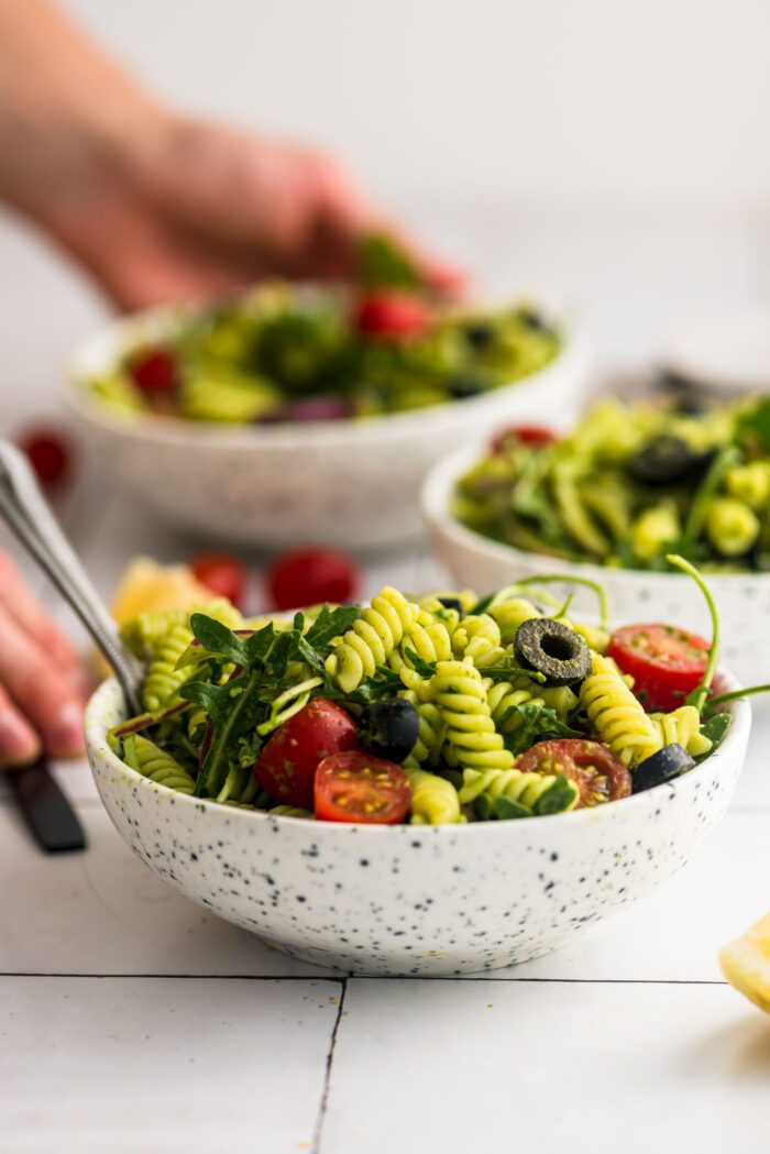 Bowl of cold pesto pasta salad with tomato, arugula, olives and red onion. Hands are in the background using a fork in another bowl of salad.