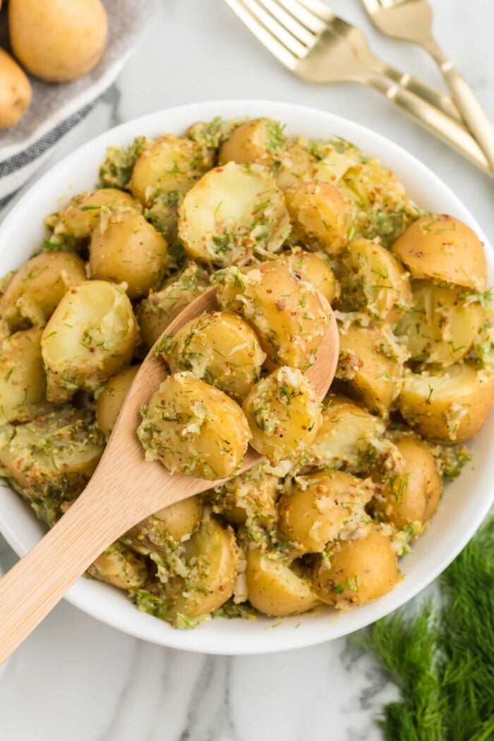 Wooden spoon lifting a few potatoes from a large bowl of dill potato salad.