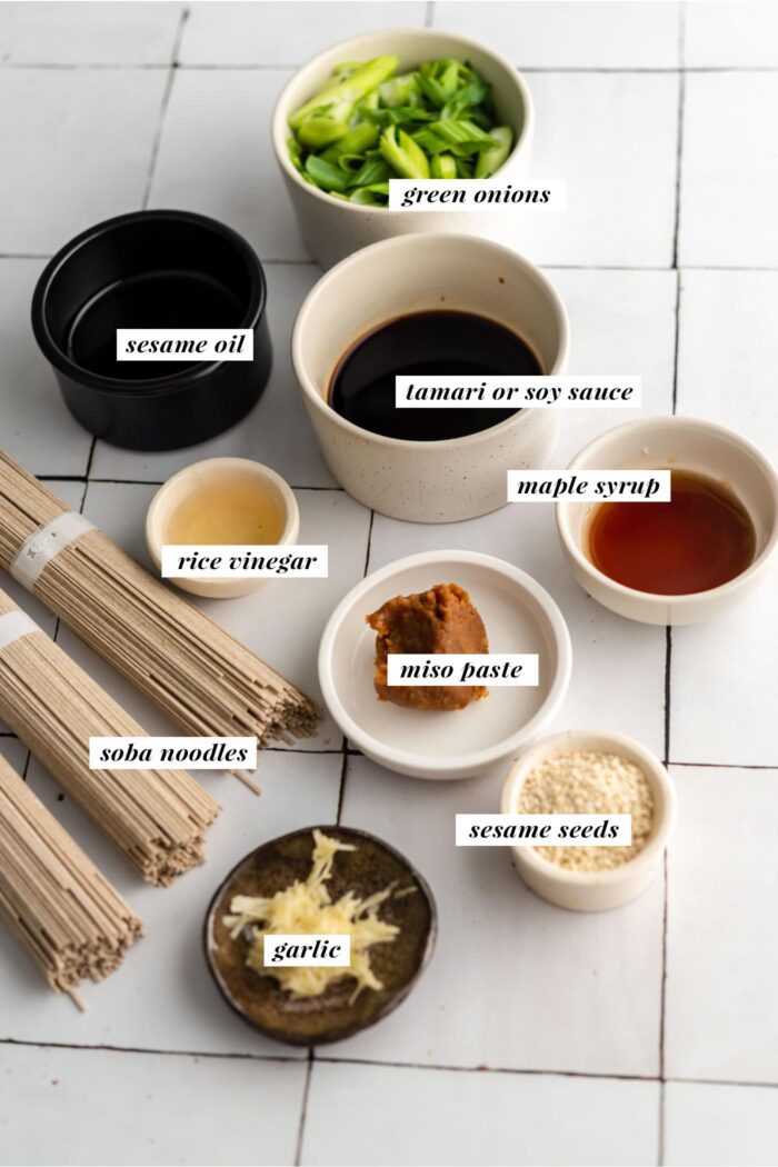 All the ingredients gathered for making a hot or cold sesame soba noodle salad with green onions. Each ingredient is labelled with text describing it.