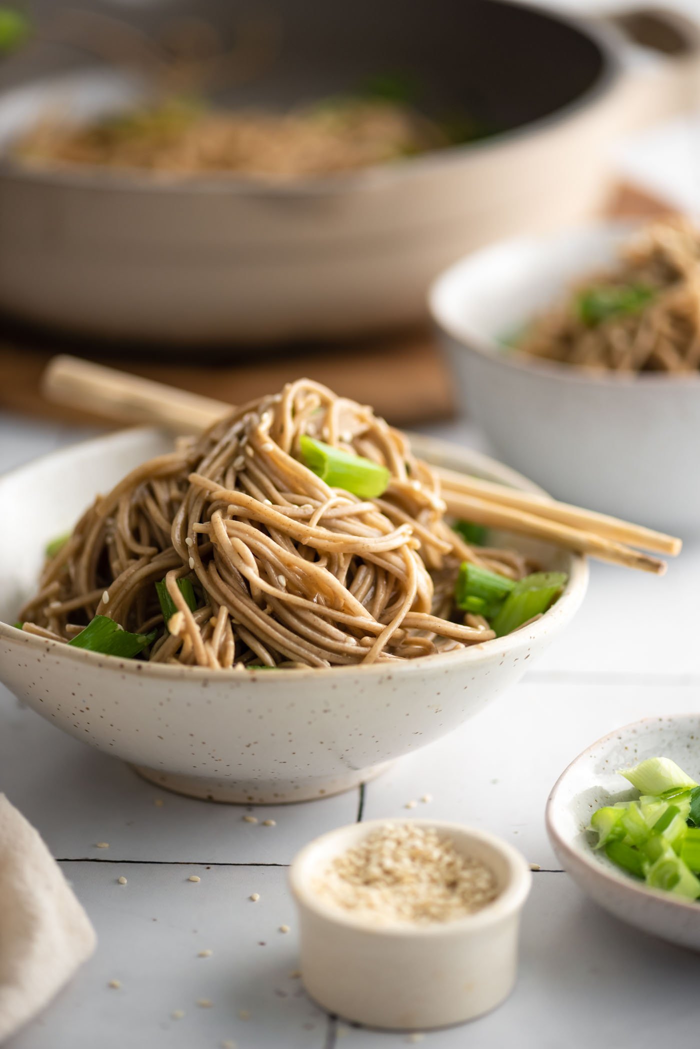 Bowl of sesame buckwheat soba noodle salad with green onions, topped with sesame seeds. A pair of chopsticks rests on the side of the bowl.