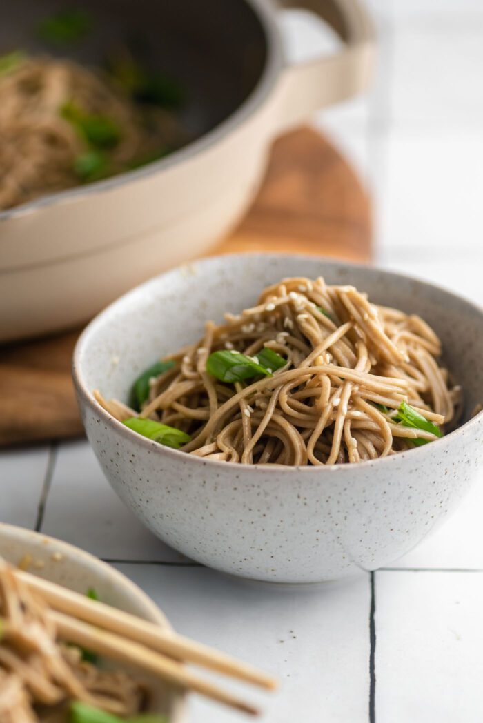 Small bowl of soba noodles with green onion in sesame dressing. A large pan of noodles is in the background and another bowl of noodles is just out of frame in the foreground.
