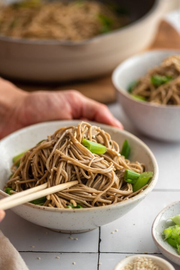 Two hands using chopsticks in a bowl of cold sesame noodle salad with green onion and sesame seeds.