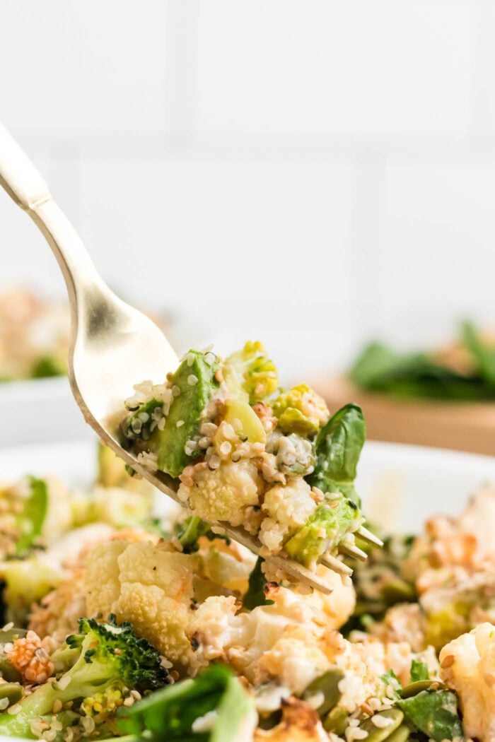 Fork with a piece of cauliflower, seeds and avocado on it. It's being held over a bowl of brocoli cauliflower salad.
