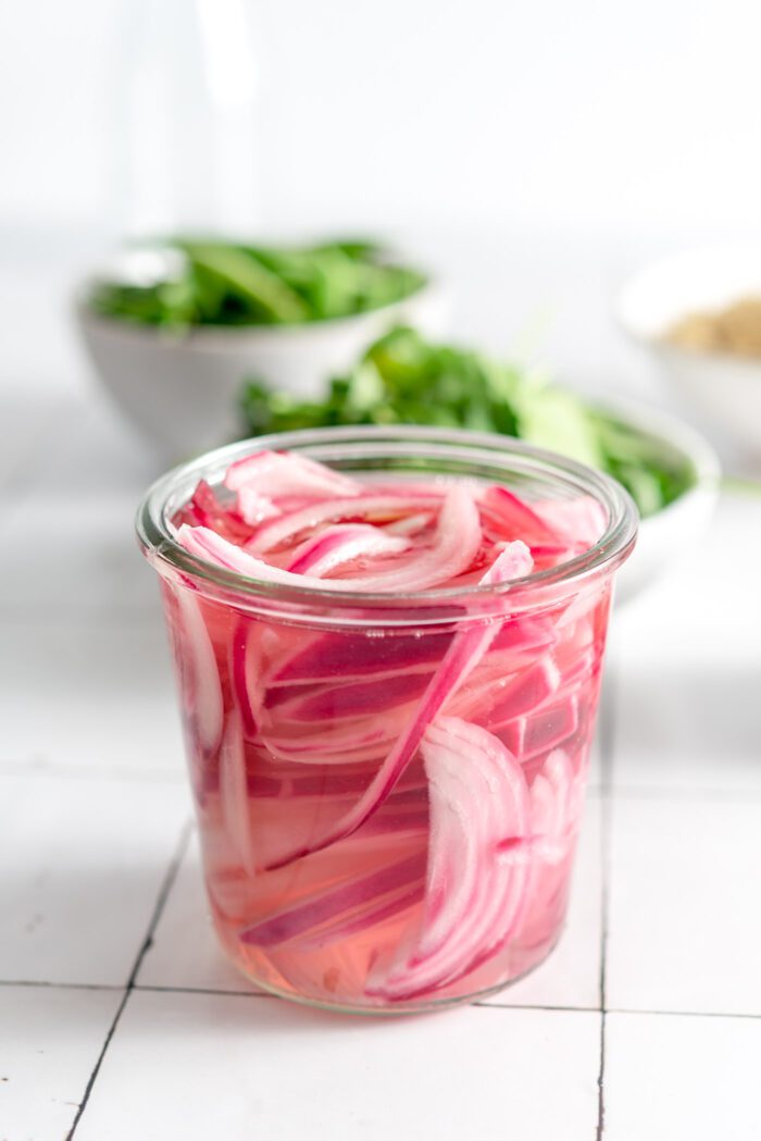 A jar of pickled red onions with two bowls of fresh greens in the background.