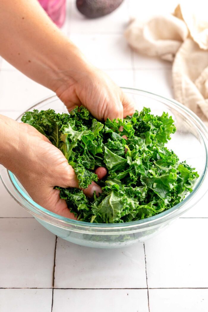 Two hands massaging chopped kale in a glass mixing bowl.