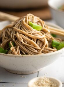 Bowl of sesame buckwheat soba noodle salad with green onions, topped with sesame seeds. A pair of chopsticks rests on the side of the bowl.