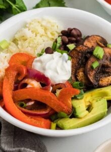 Easy veggie fajita bowls with cooked bell peppers and onions, avocado, rice, sour cream, black beans and marinated cooked mushrooms.