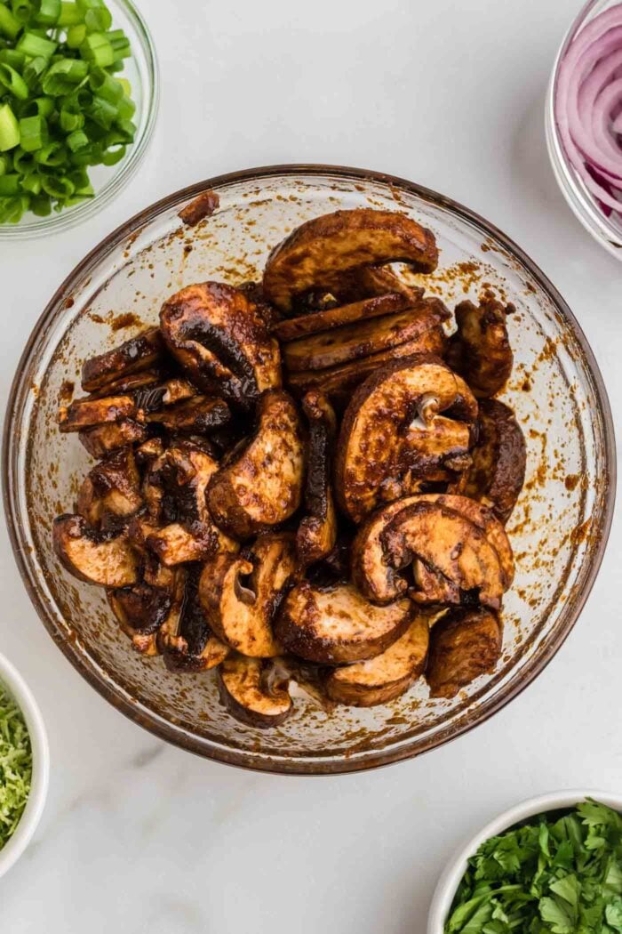 Sliced mushrooms marinated in fajita spices, soy sauce and balsamic vinegar in a glass bowl.