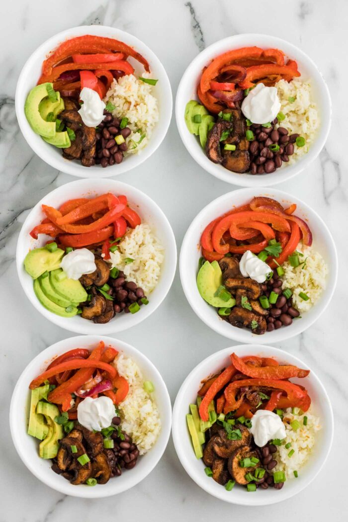 6 vegetarian fajita bowls with rice, black beans, mushrooms and cooked peppers and onions. Each is topped with sliced avocado and cilantro..