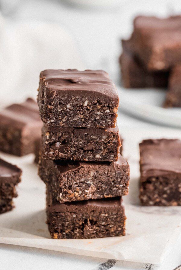 Stack of 4 no-bake hemp see brownies topped with chocolate frosting with more brownies scattered in the background.