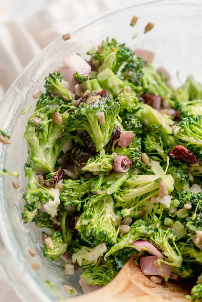 Close up of vegan broccoli salad with mayo dressing and cranberries and sunflower seeds in a large mixing bowl with a wooden spoon resting in the bowl.