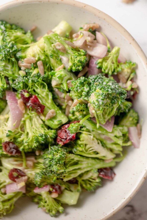 Close up of vegan broccoli salad with mayo dressing and cranberries and sunflower seeds in a small ceramic serving bowl.