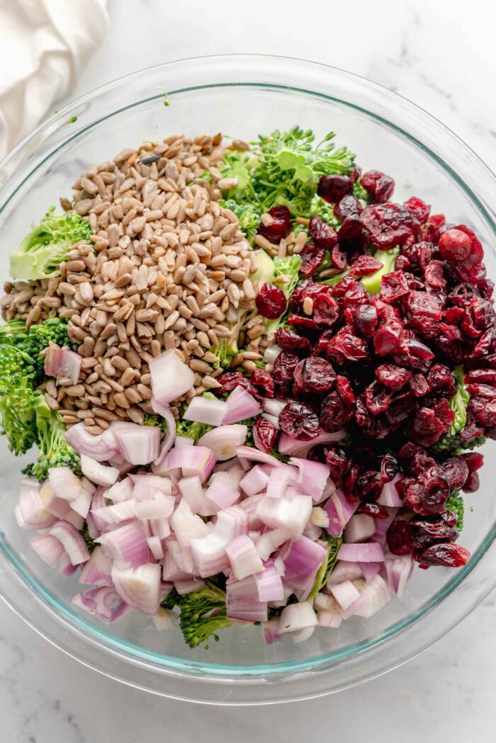 Diced red onion, dried cranberries, sunflower seeds and broccoli in a large glass mixing bowl.