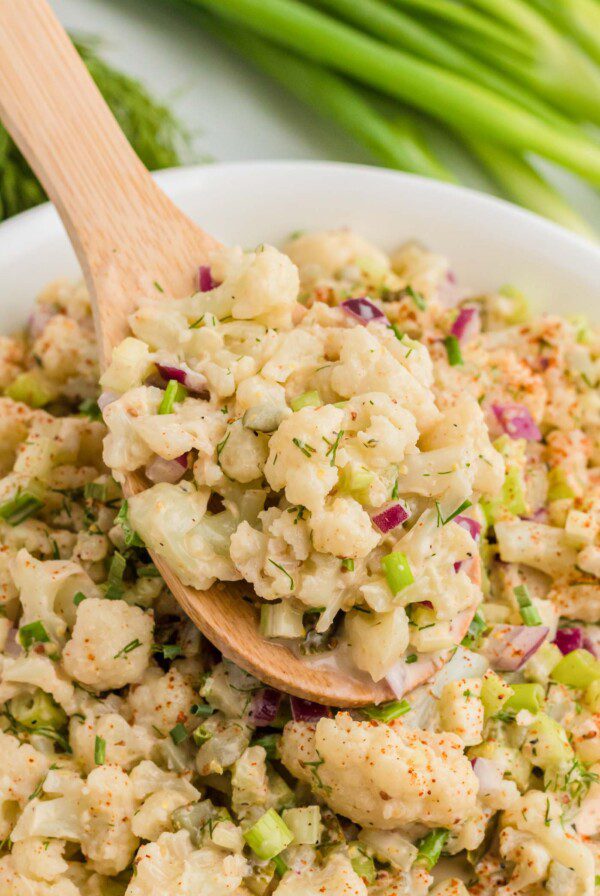 Wooden spoon scooping cauliflower mock potato salad from a large serving bowl of salad. There are some green onions in the background.