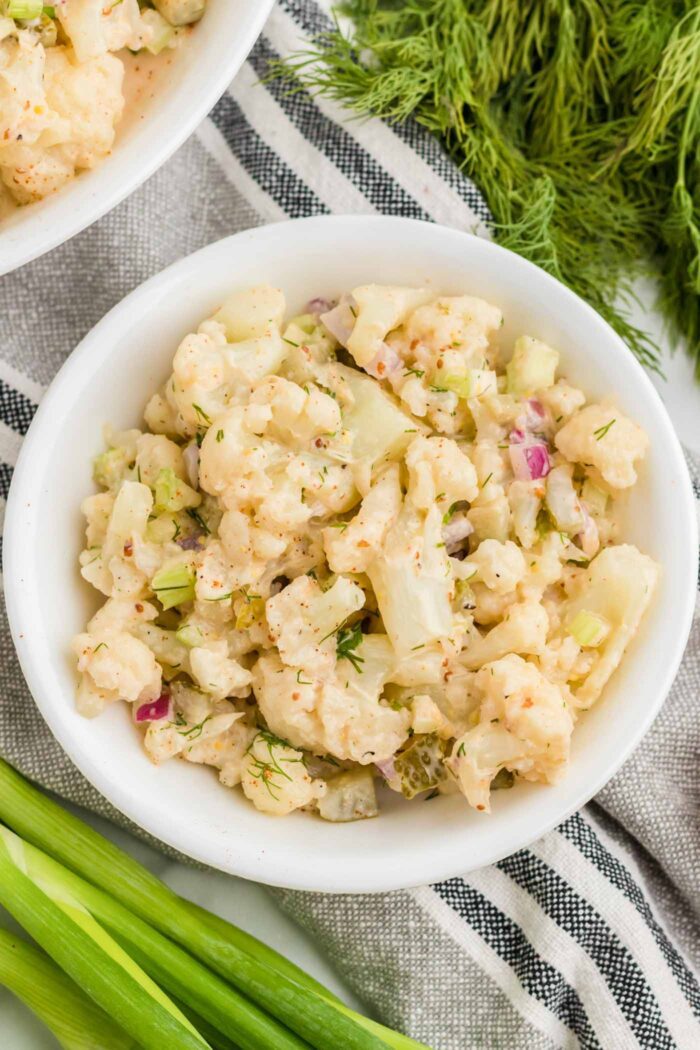 Overhead view of a bowl of cauliflower potato salad with dill pickle bits, celery, red onion and scallions. It's sprinkled with smoked paprika and there is some fresh dill and scallions resting beside the bowl.