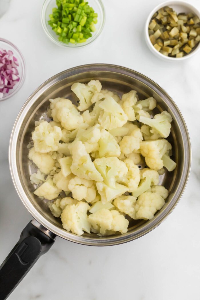 Steamed cauliflower florets in a steamer pot. There are 3 small bowls beside the pot, one with dill pickled, one with red onion and one with chopped green onion.