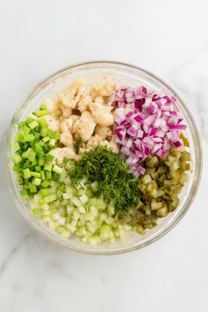 Steamed cauliflower, diced red onion, celery, dill pickle, fresh dill and scallions in a glass mixing bowl on a marble countertop.