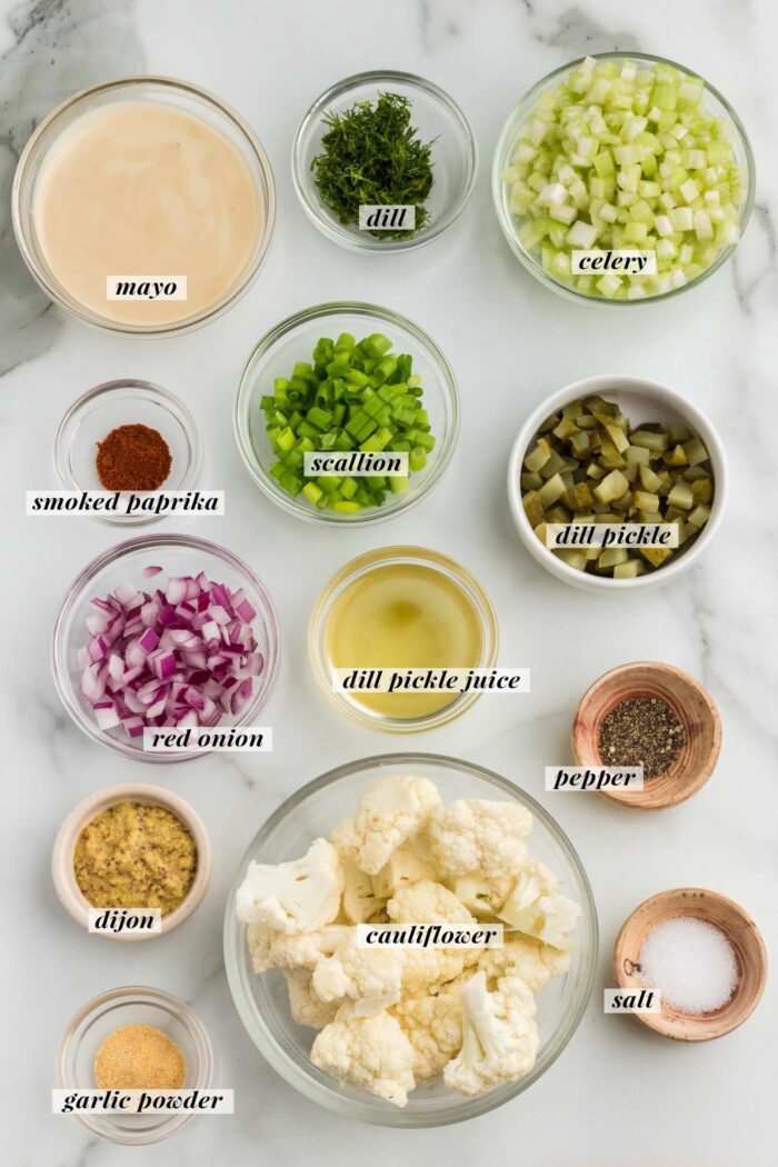 All the ingredients gathered in bowls for making a cauliflower "potato" salad with dill pickled, red onion, celery and spices. Each ingredient is labelled with text.